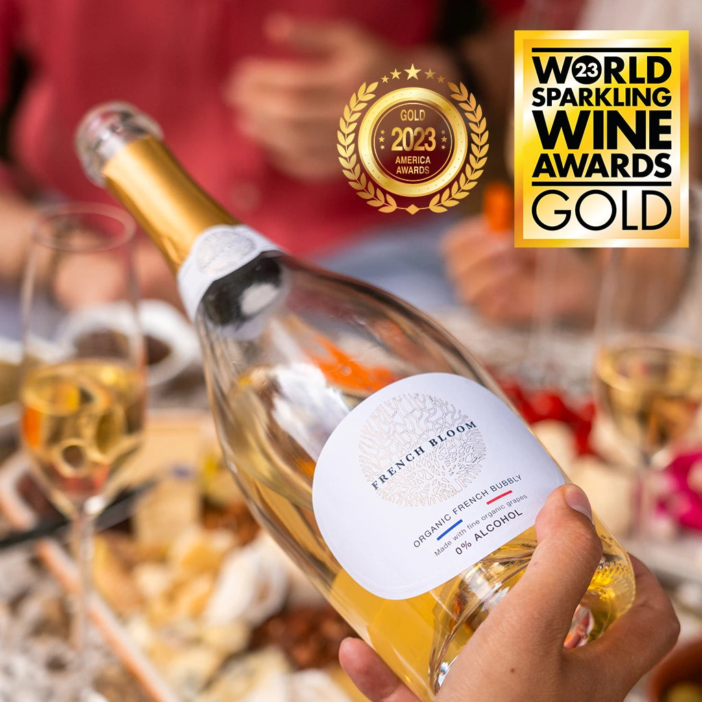 Eine Flasche French Bloom Le Blanc mit der Auszeichnung "Gold 2023 World Sparkling Wine Awards" im Vordergrund, umgeben von einem gedeckten Tisch und Gläsern mit Schaumwein.