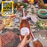 Eine Flasche French Bloom Le Rosé mit der Auszeichnung "Gold 2023 World Sparkling Wine Awards", gehalten über einem gedeckten Tisch mit sommerlichem Geschirr und Speisen.