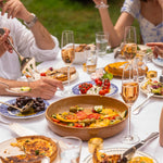 Ein geselliger Sommertisch im Freien, gedeckt mit frischen Speisen, Oliven, Tomaten und French Bloom Le Rosé in Gläsern, während Menschen miteinander essen und anstoßen.