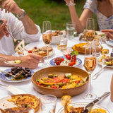 Ein geselliger Sommertisch im Freien, gedeckt mit frischen Speisen, Oliven, Tomaten und French Bloom Le Rosé in Gläsern, während Menschen miteinander essen und anstoßen.