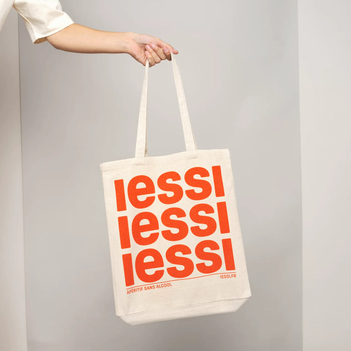 IESSI Tote Bag: heller Stoffbeutel mit dreifachem orangem IESSI-Schriftzug, an der Hand gehalten