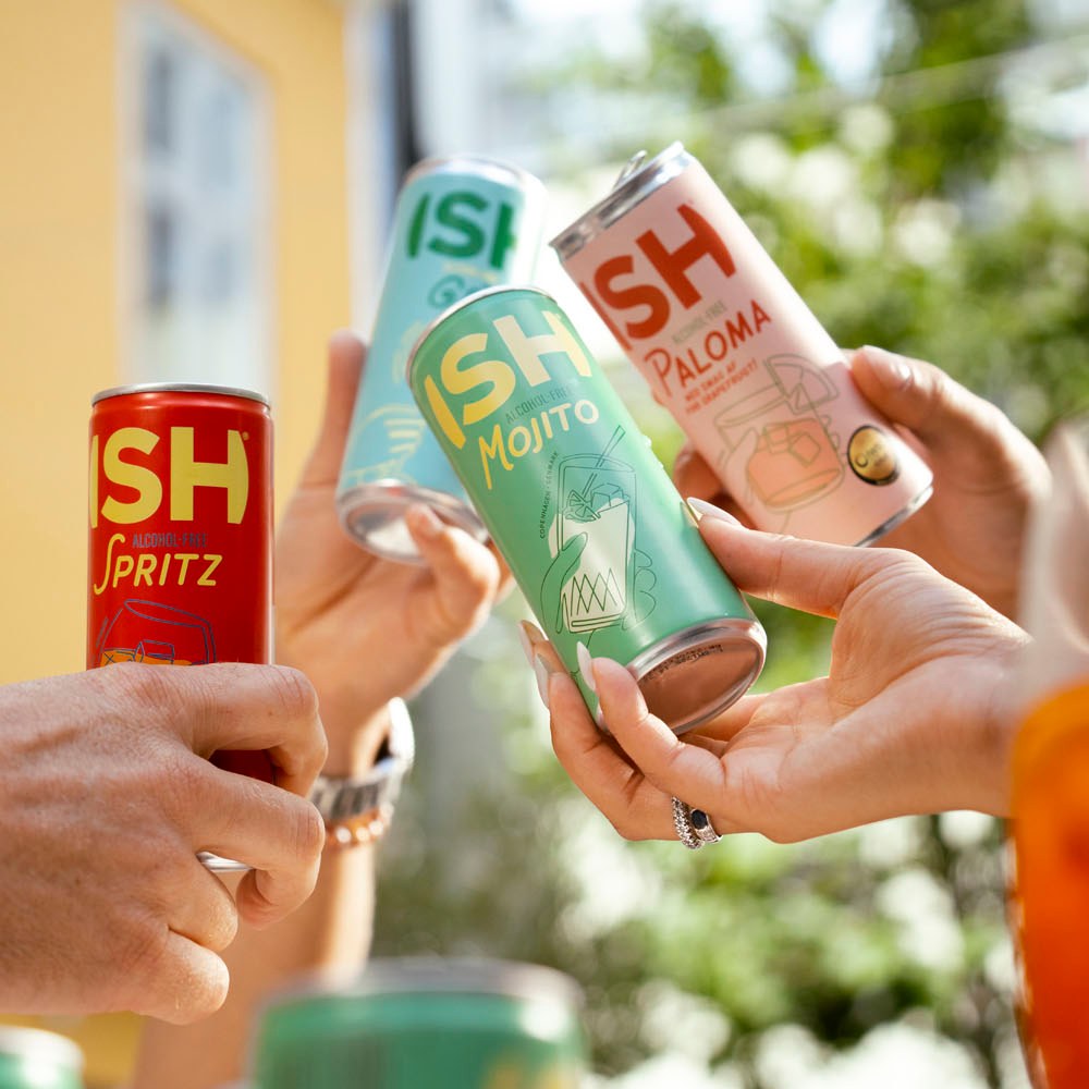 Hände stoßen im Sonnenlicht mit Dosen ISH Spritz, Mojito und Paloma an
